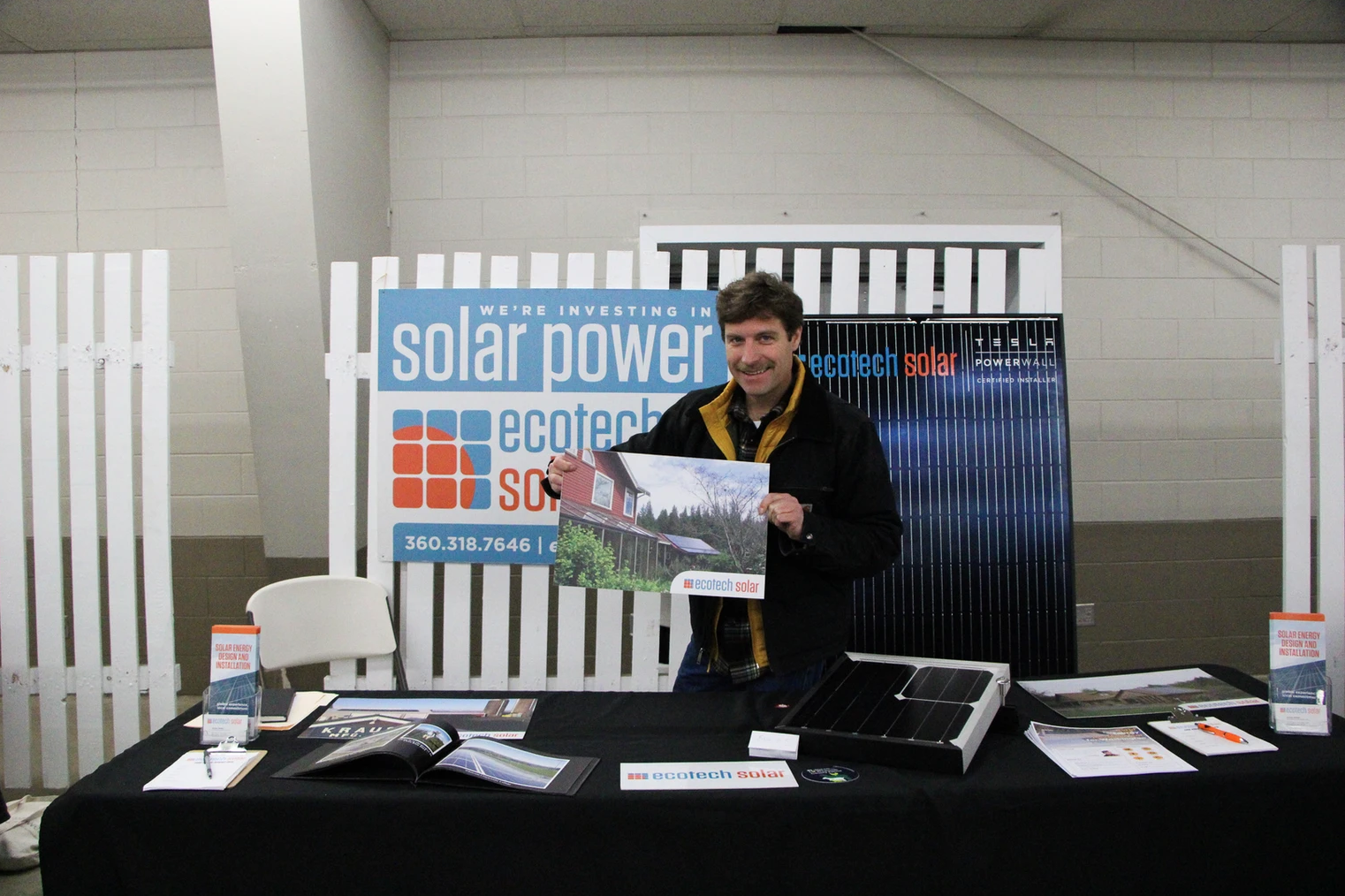 A man holding a photo at a solar power booth with promotional materials on a table.
