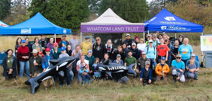 A group of people posing in front of "WHATCOM LAND TRUST" and other tents, holding inflatable orcas.