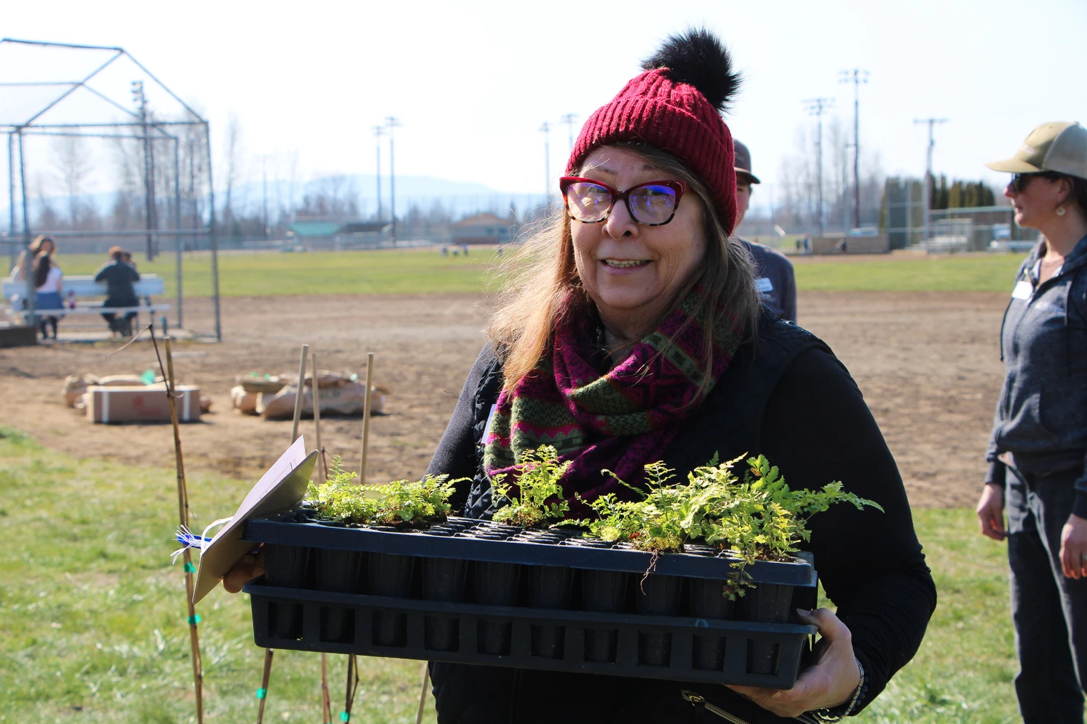 A person in a red beanie holds a tray of small plants in a field, with others nearby.