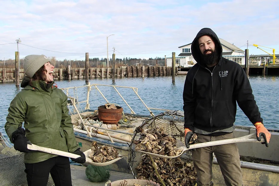 Two people are smiling on a boat, holding shovels filled with oysters, in a harbor setting.