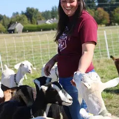 A person smiling in a field surrounded by goats.
