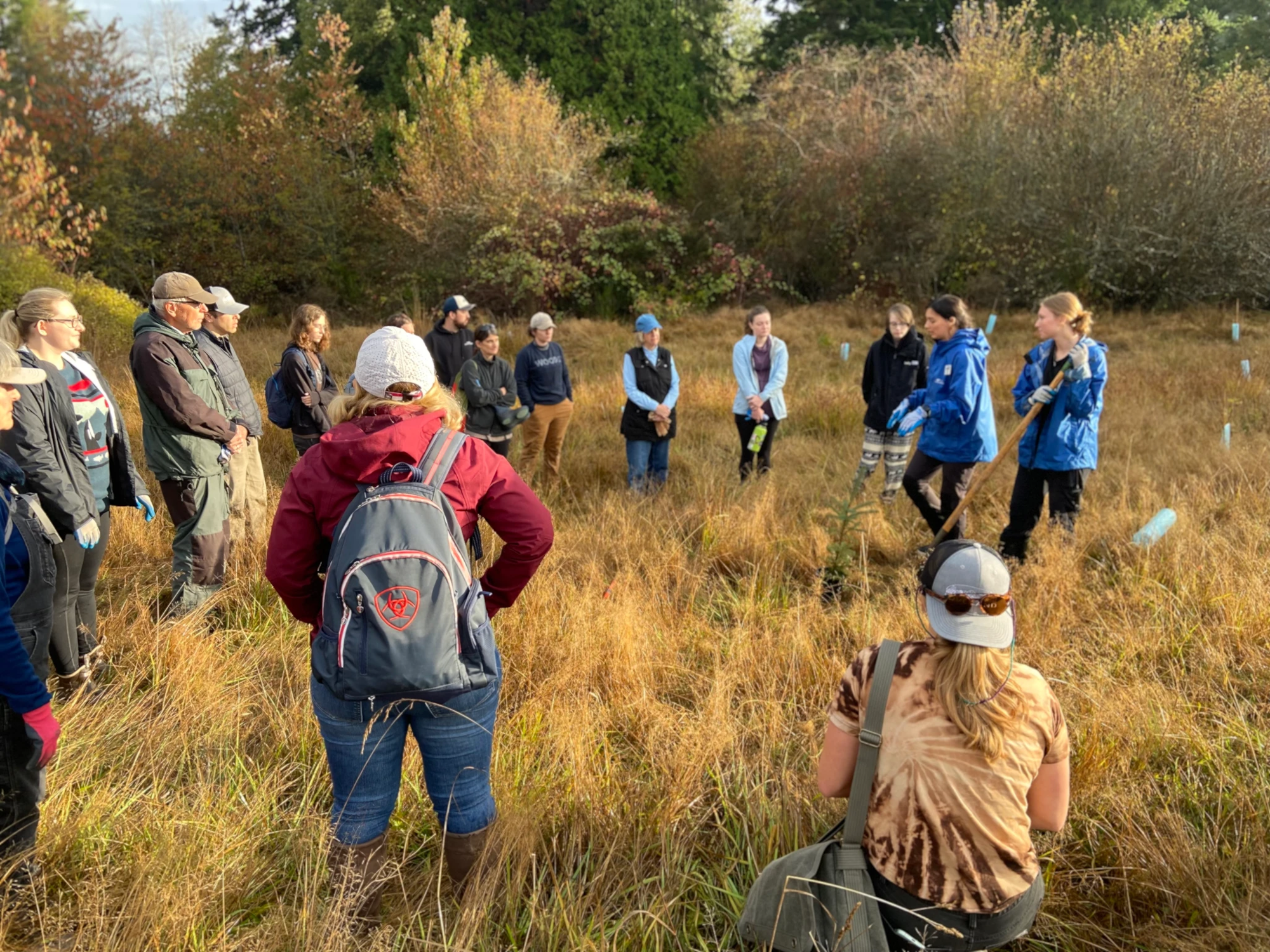 A group of people in an outdoor field, some holding tools, appearing to engage in a discussion or activity.