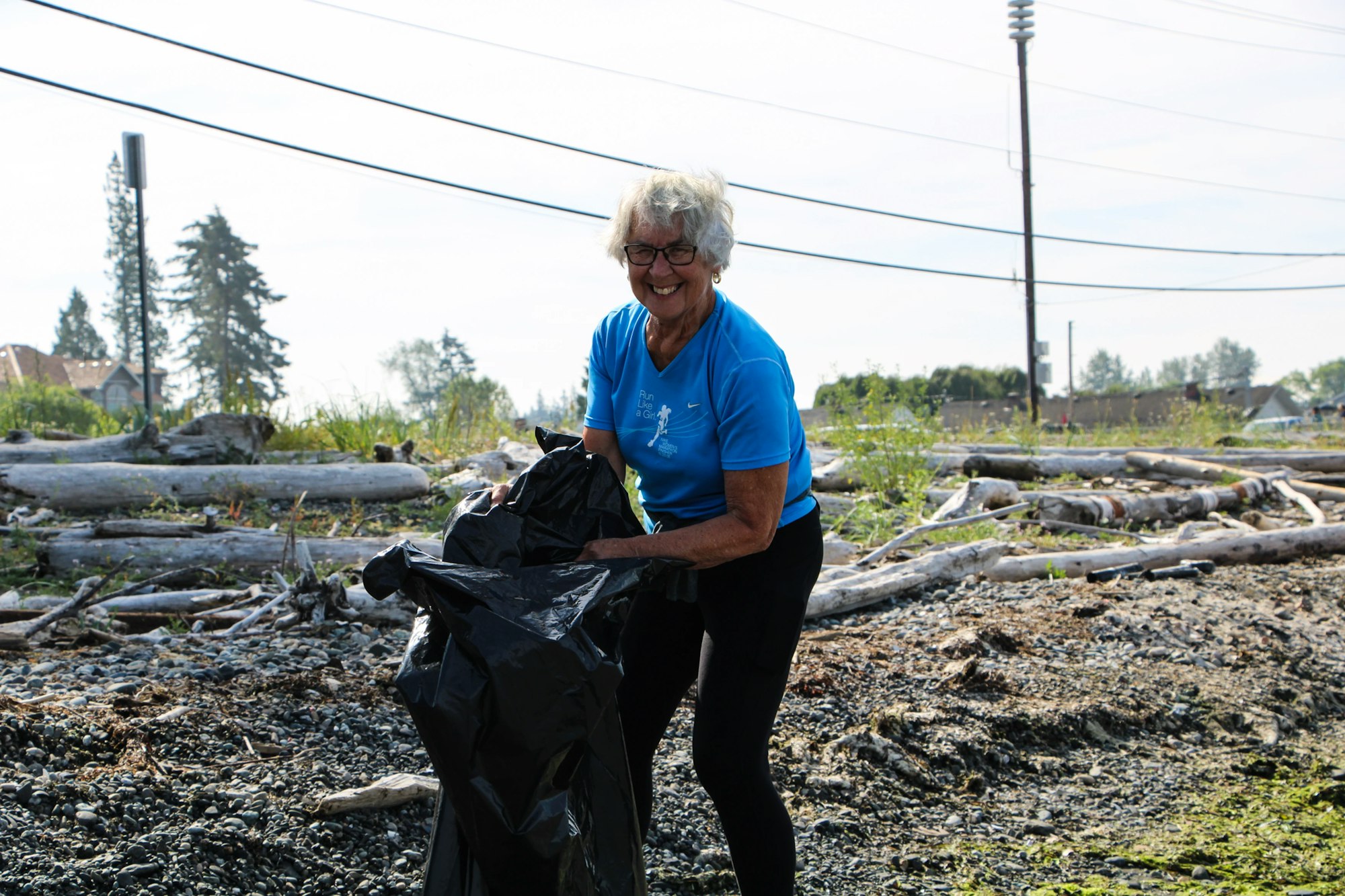 A woman in a blue shirt is cleaning up debris on a rocky area, holding a black garbage bag.