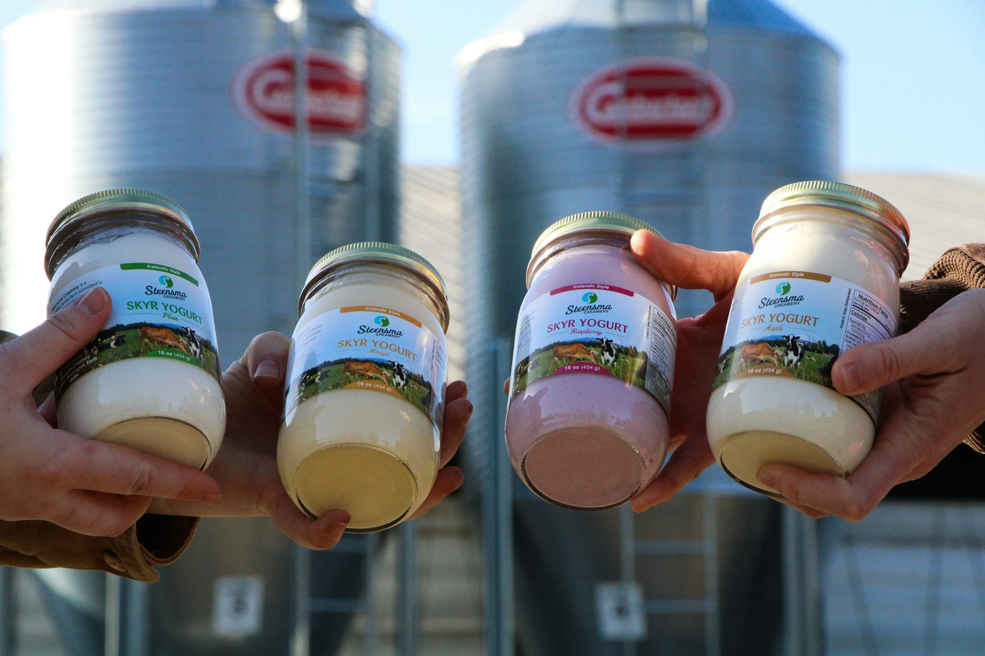 Four hands holding jars of Steensma Skyr Yogurt with grain silos in the background.
