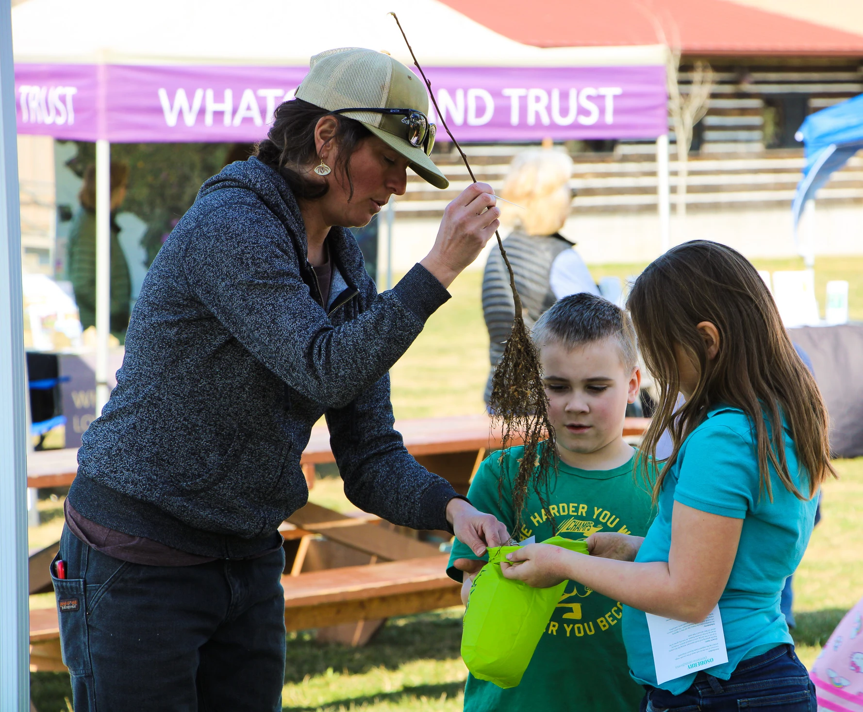 A woman shows a plant with roots to two children outdoors. They're near a "LAND TRUST" sign.