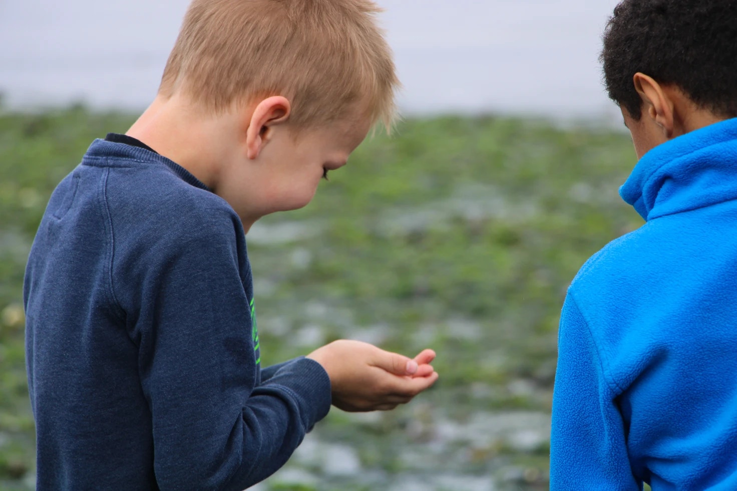 Two children outside, one examining something in their hands, wearing blue jackets.