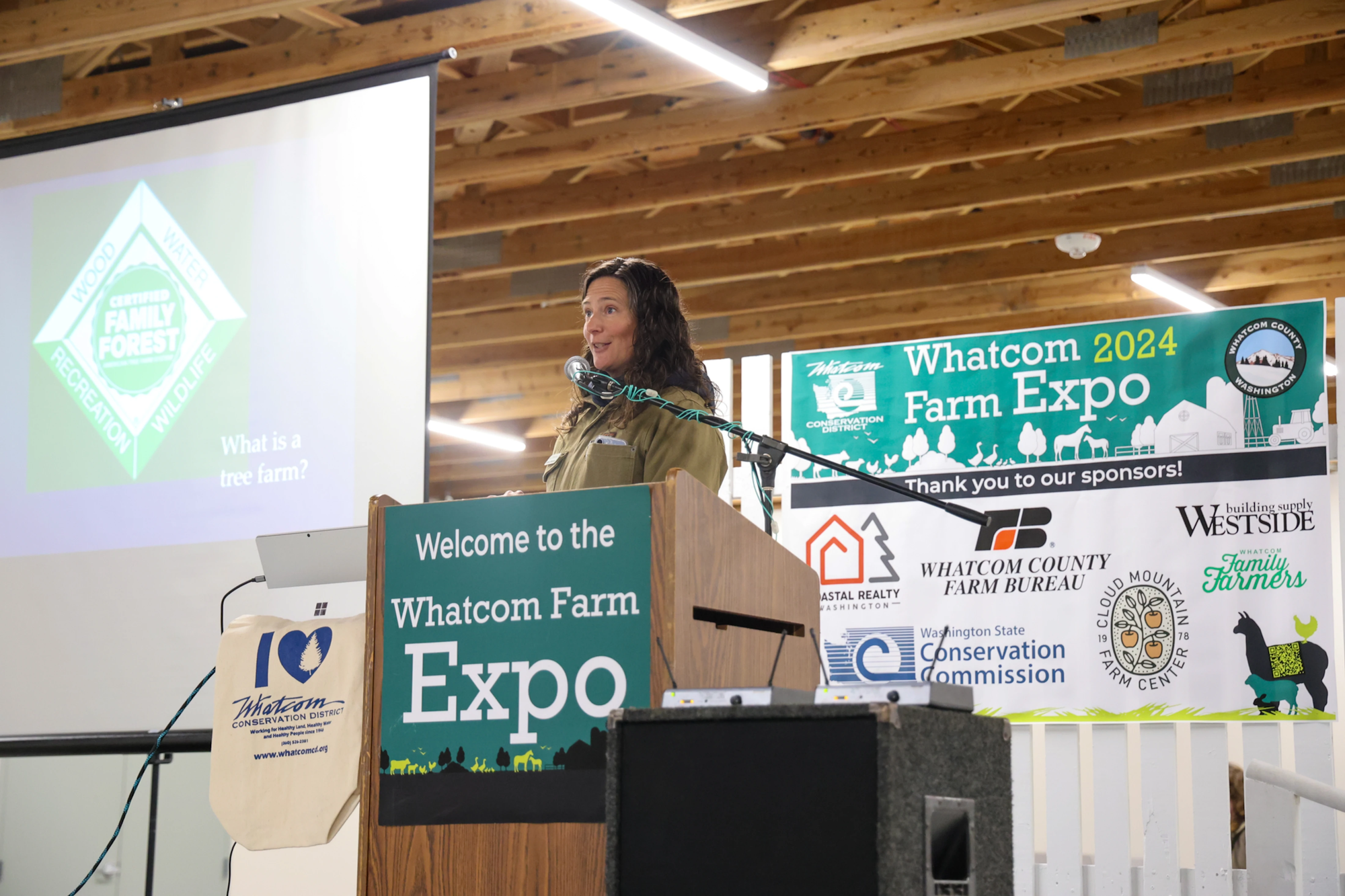 A woman speaks at the 2024 Whatcom Farm Expo. A sign behind her thanks sponsors. A screen reads "What is a tree farm?"
