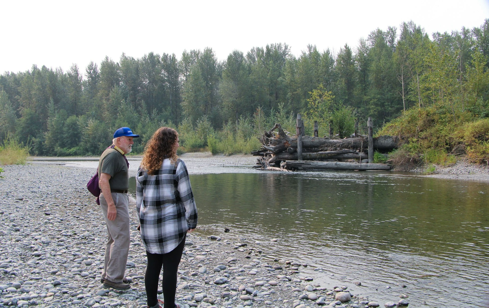 Two people stand on a rocky riverbank, observing a log structure in a tranquil river, surrounded by trees.
