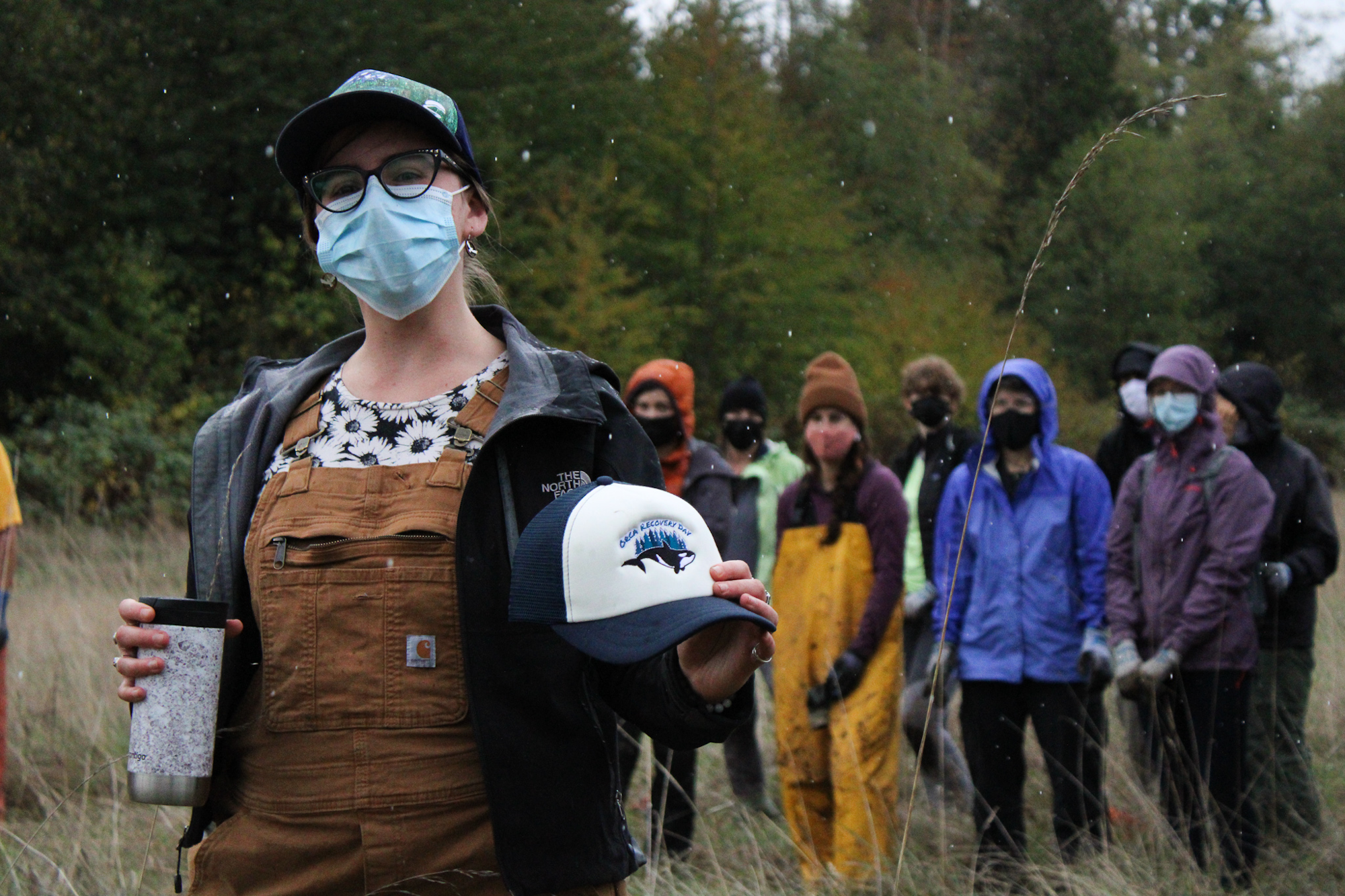 A person in a mask holds a hat outdoors, with a group of masked people in the background.