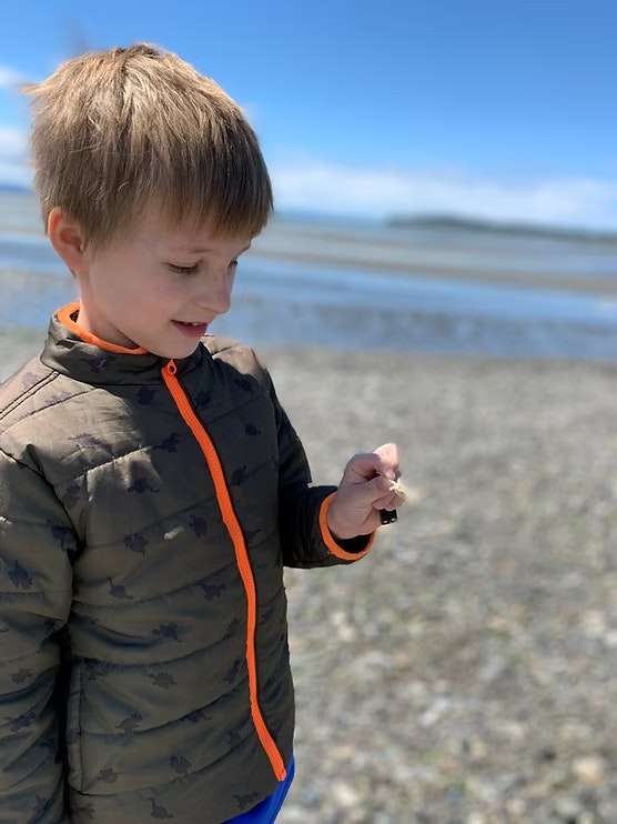 A child on a rocky beach holding something small, wearing a jacket with an orange zipper, under a blue sky.