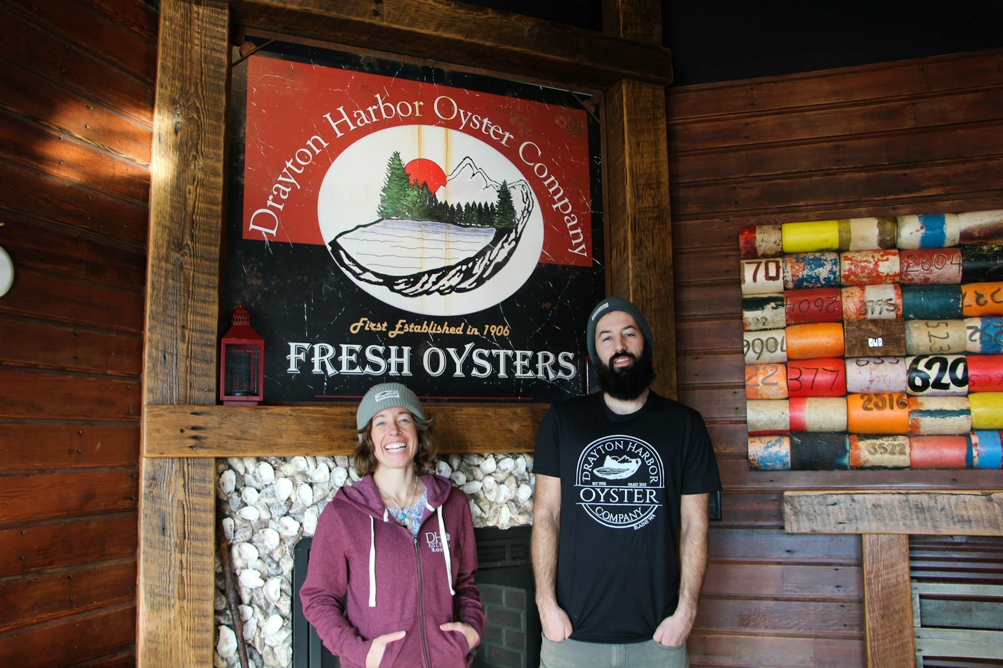 Two people in front of a "Drayton Harbor Oyster Co." sign, promoting fresh oysters.