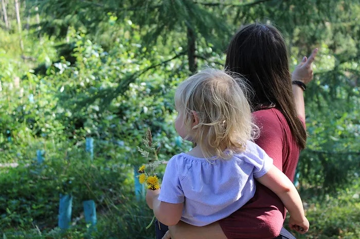 An adult and child with flowers, back to camera, surrounded by greenery.