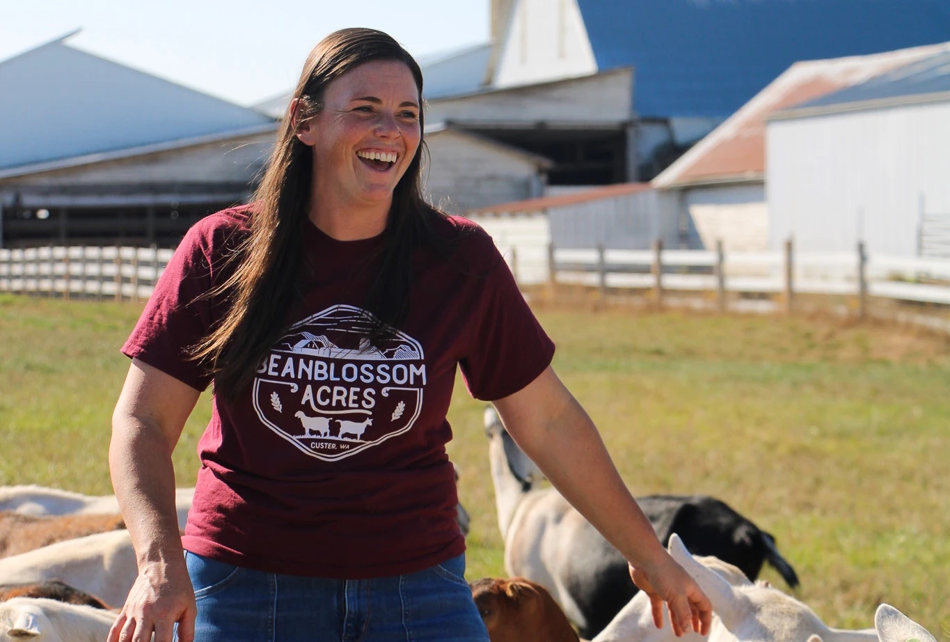 A person smiling on a farm, wearing a "Beanblossom Acres" t-shirt, surrounded by goats.