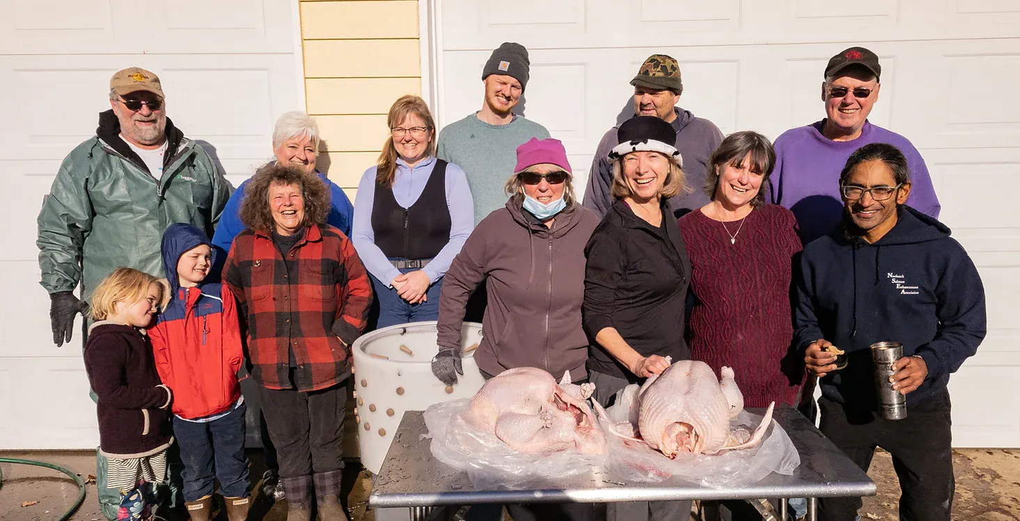 Group of people smiling outdoors with two raw turkeys on a table.