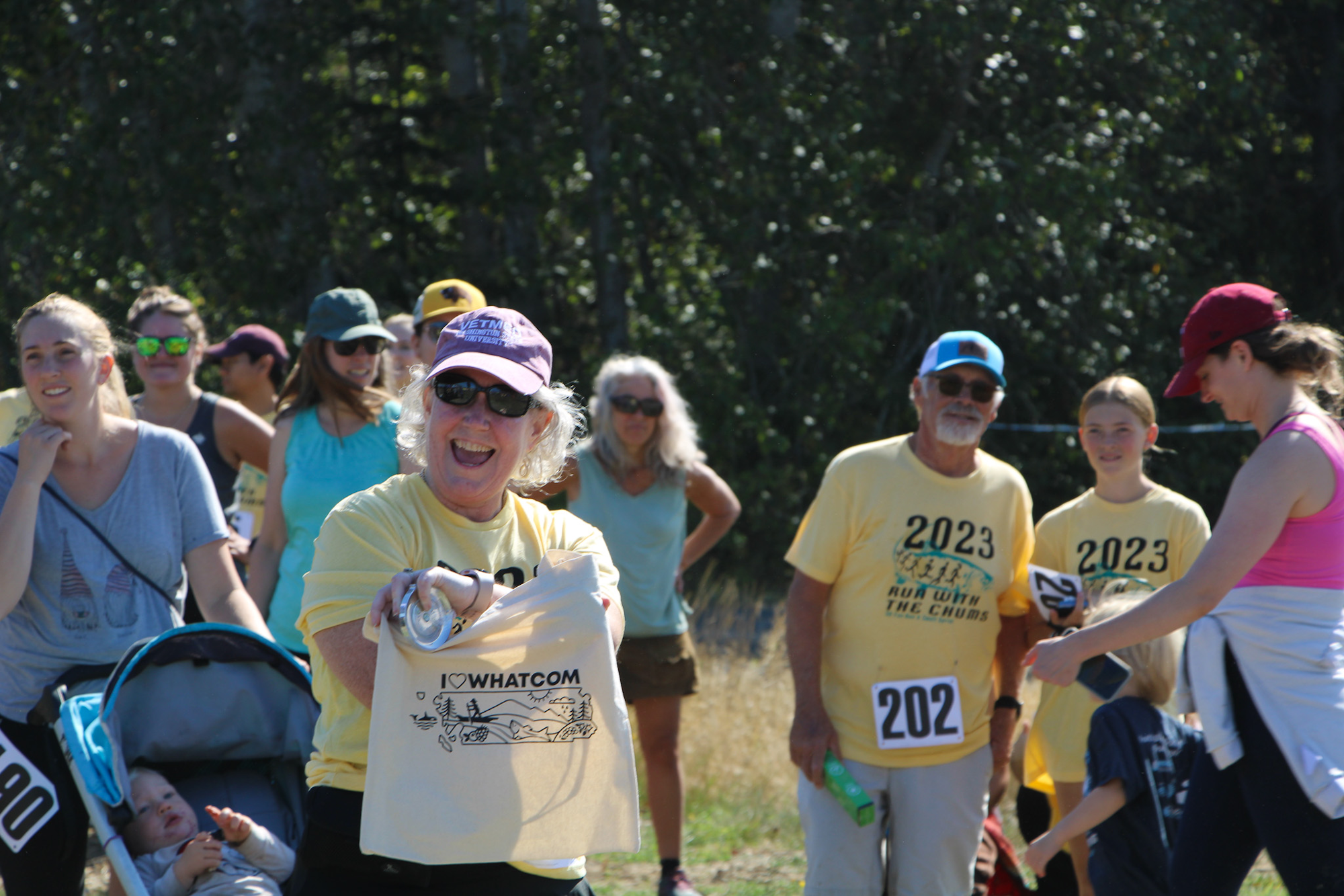 A group of people outdoors, some wearing yellow "2023 Run with the Chums" shirts, one holding an "I Love Whatcom" bag.