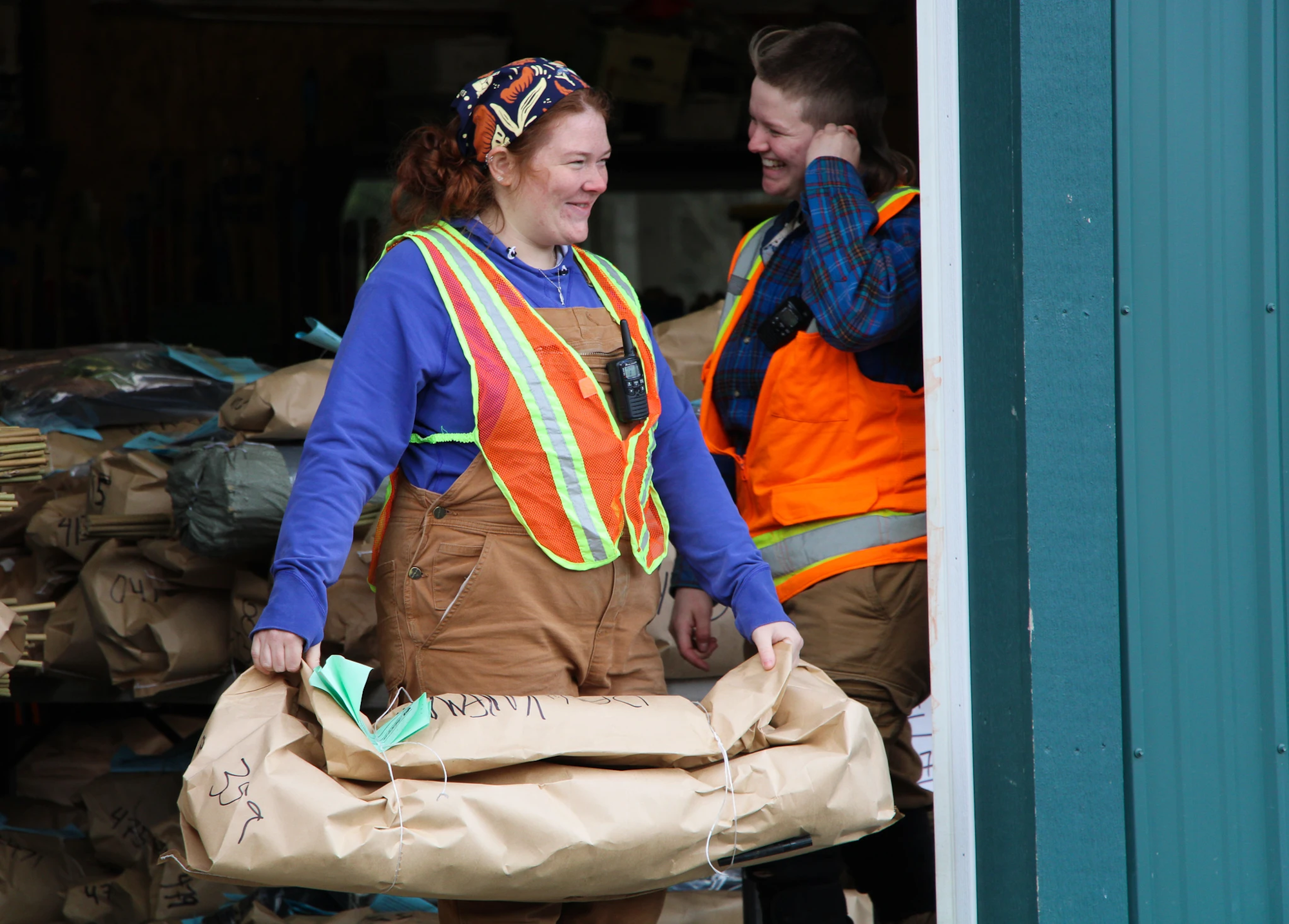Two people wearing safety vests and work clothes, one carrying a large paper-wrapped package.