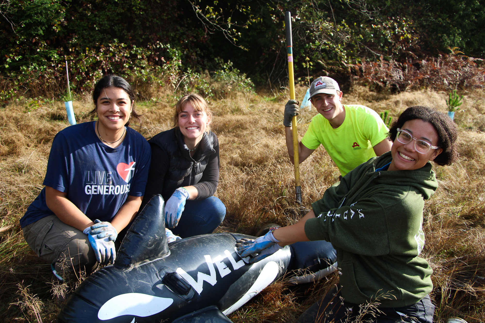 Four people smiling outdoors, holding a black inflatable with “WEL” on it, against a grassy background.