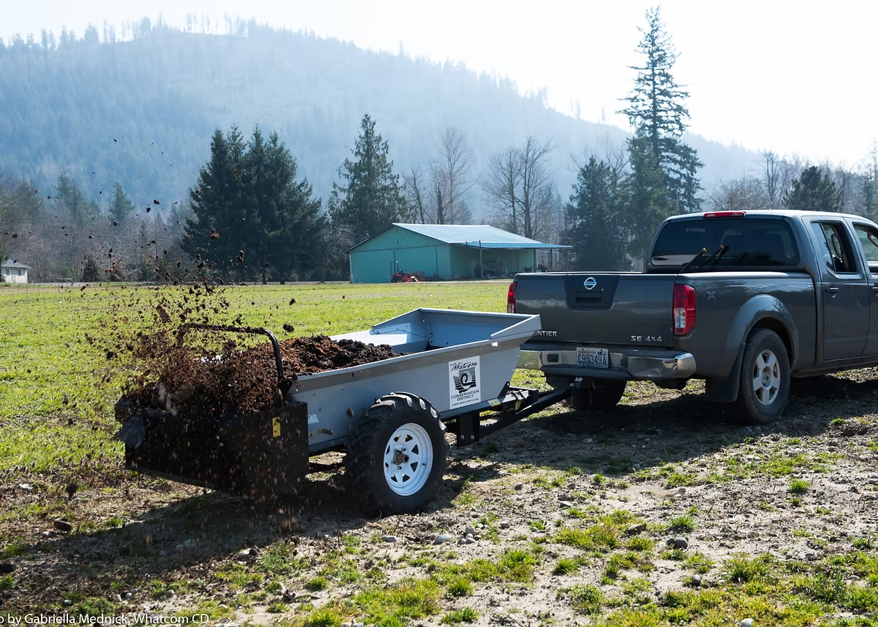 A truck tows a trailer, spreading manure on a grassy field near a barn and trees.