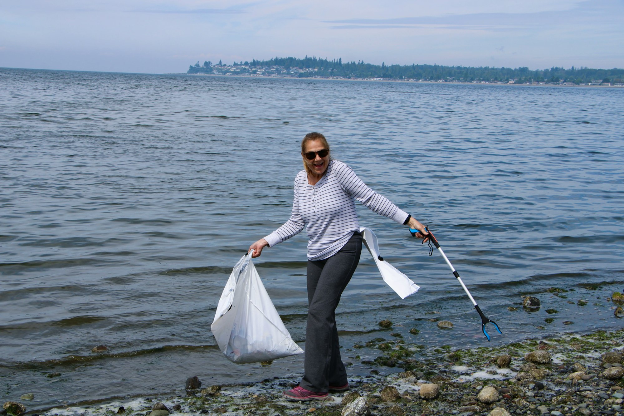 A person cleaning a beach, holding a trash bag and a grabber tool, smiling by the water.