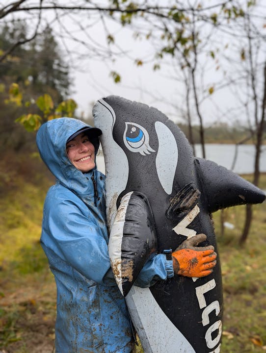 Person in a blue raincoat hugging an inflatable orca, smiling, in a rainy outdoor setting.