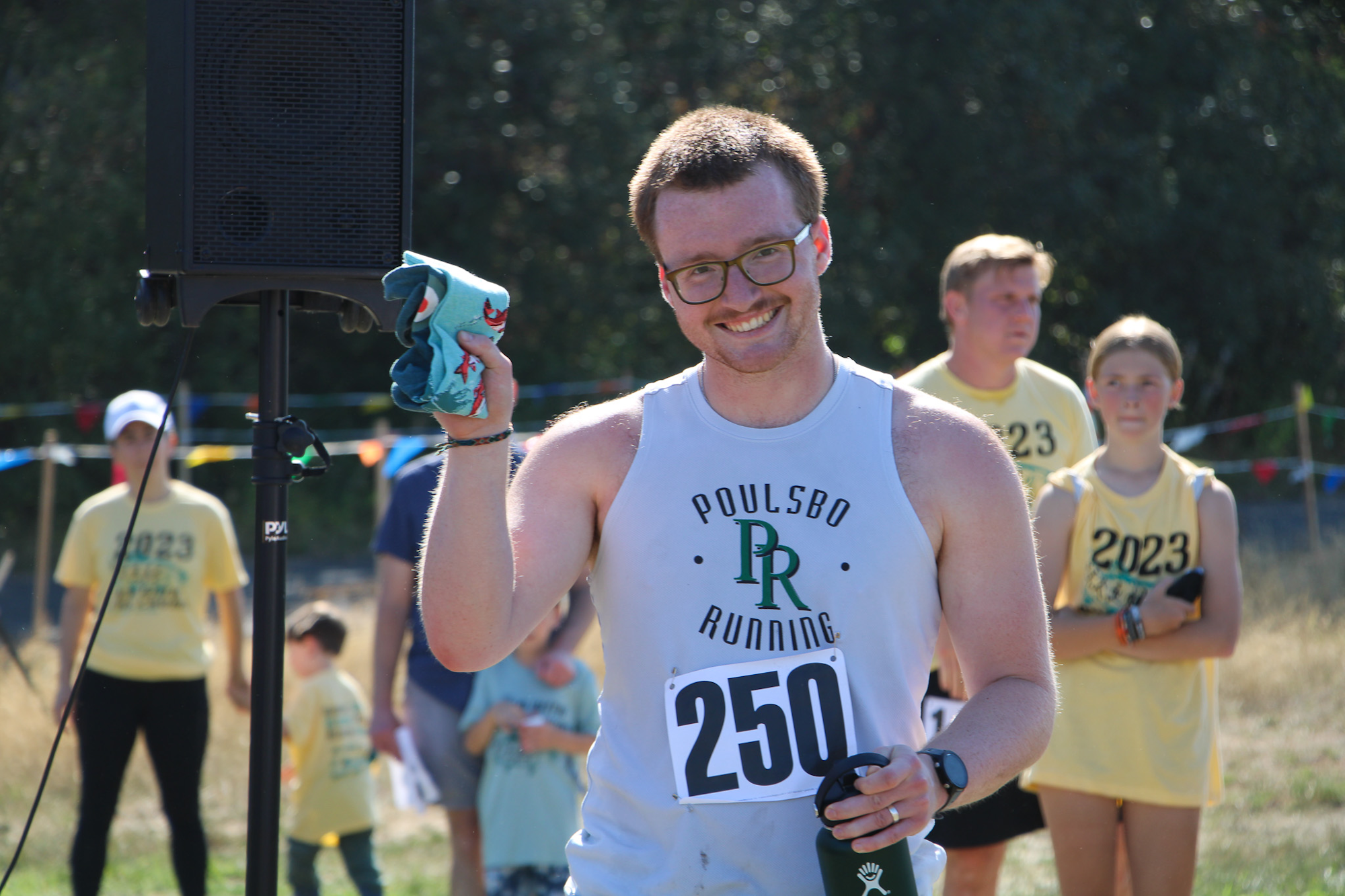 A smiling runner holding a towel and bottle, wearing a "Poulsbo Running" shirt with the number 250, and others in the background.