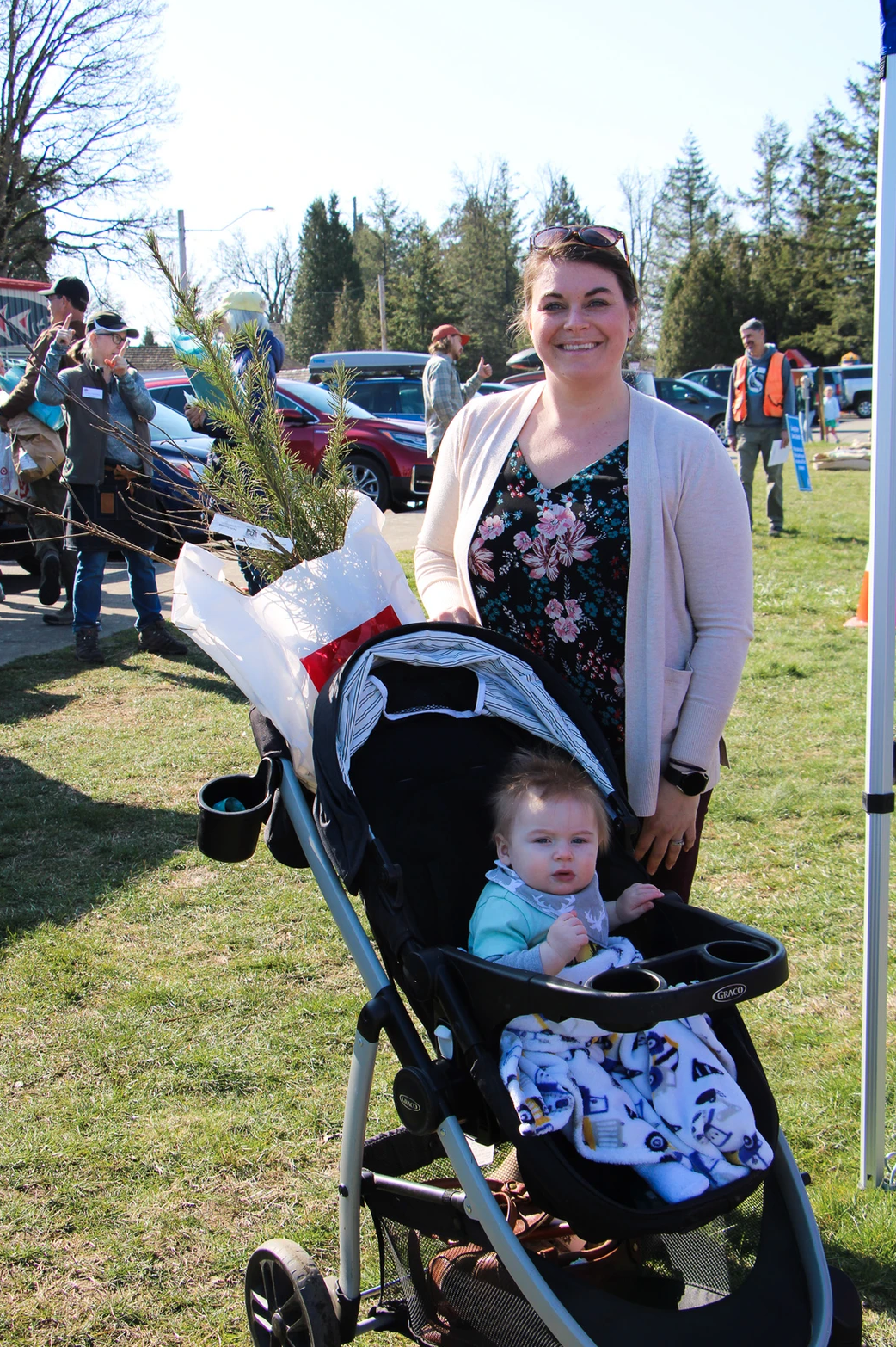 A woman with a stroller holding a baby and a plant in an outdoor setting with trees and people in the background.