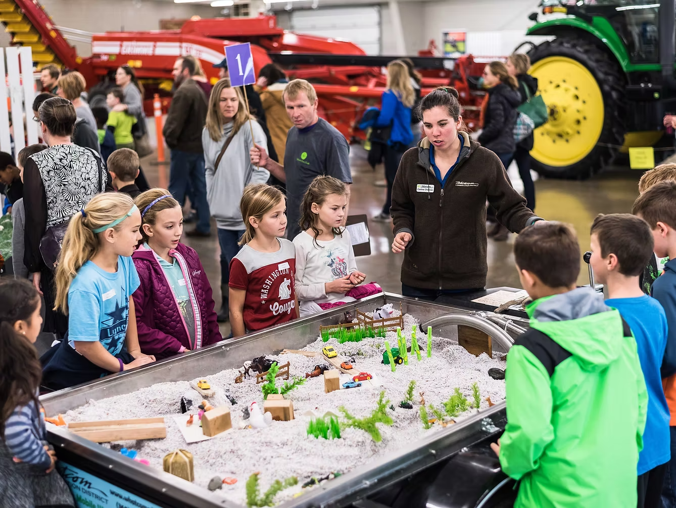 A woman explains a model farm display to a group of children at an indoor event with agricultural machinery in the background.