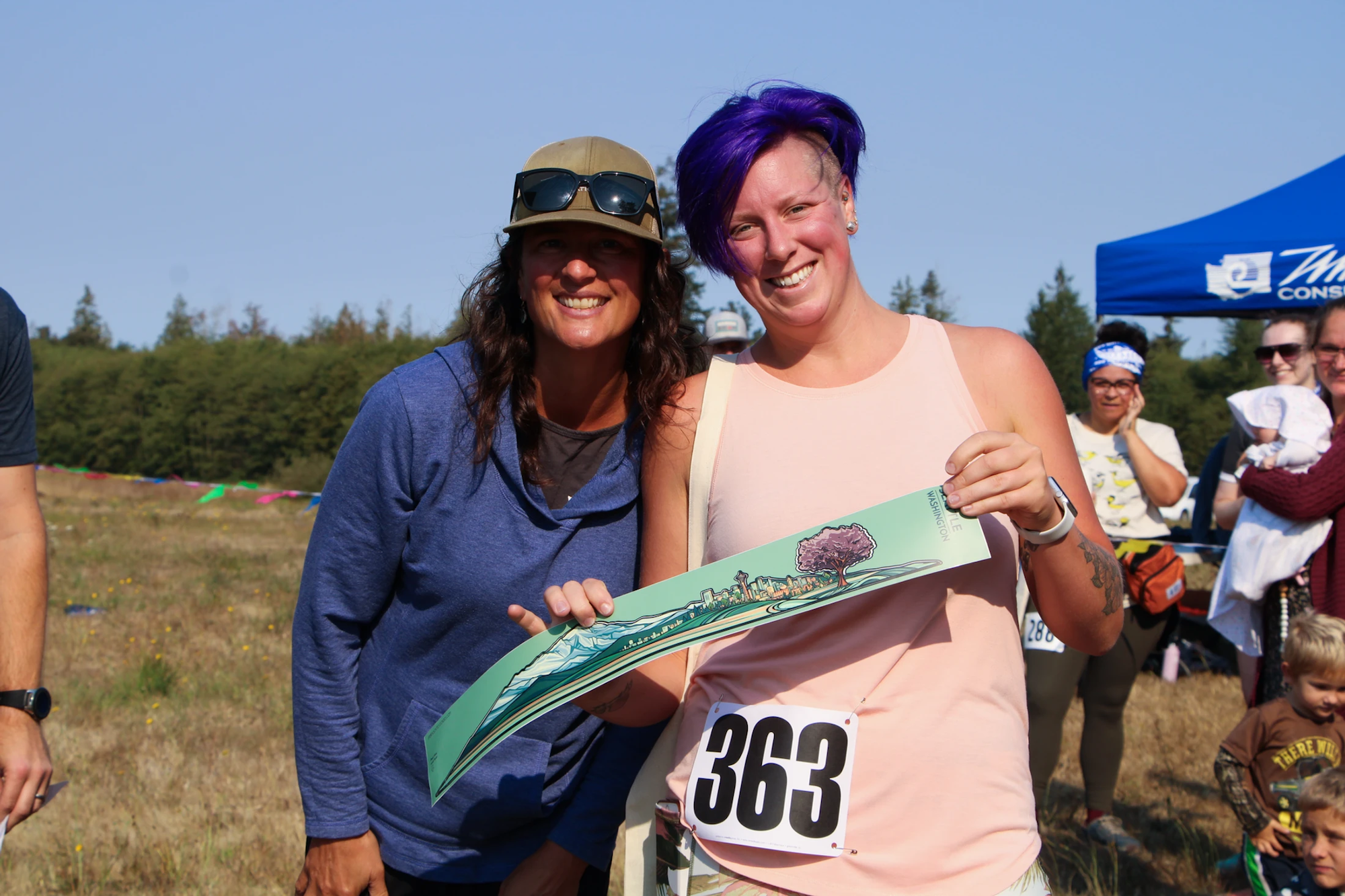 Two people smiling, one holding a colorful certificate. Others in the background outdoors.