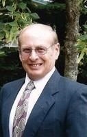 A smiling person in a suit and tie, standing outside with trees in the background.
