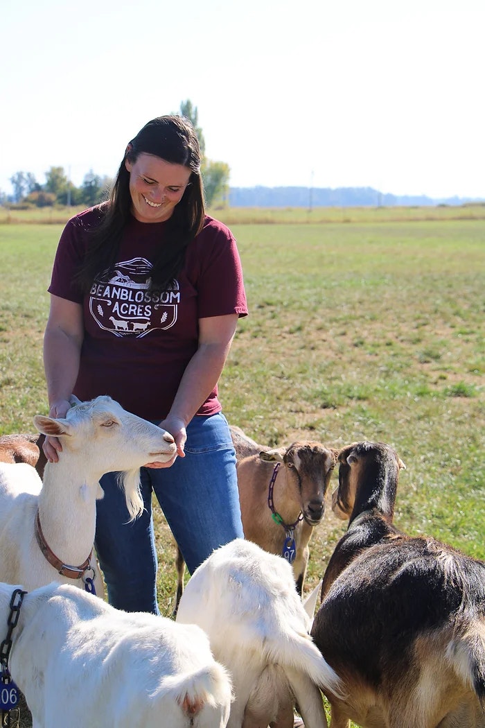 A person smiling, surrounded by goats in a field, wearing a maroon shirt with "Beanblossom Acres" text.