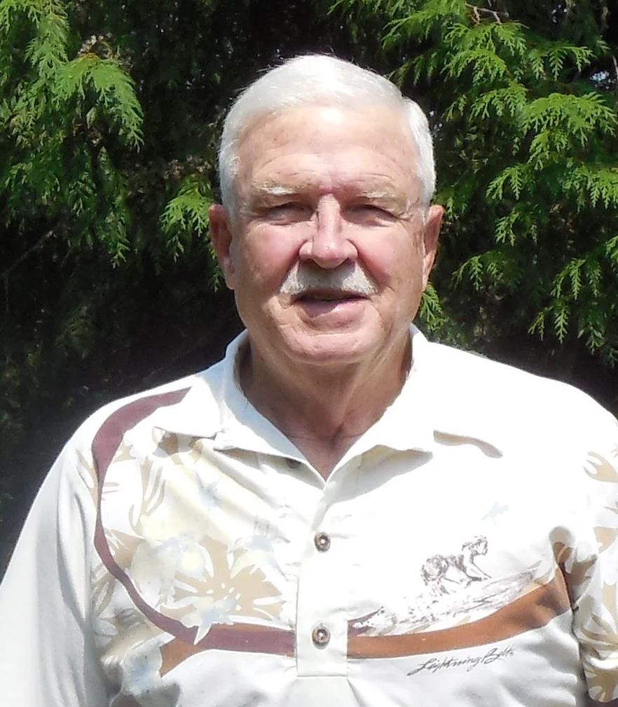 A man with white hair and a mustache, wearing a patterned shirt, stands outdoors with greenery in the background.