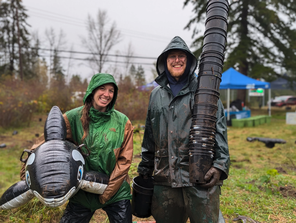 Two people in rain gear smiling; one holds an inflatable orca, the other a stack of plant pots. Outdoors, overcast weather.