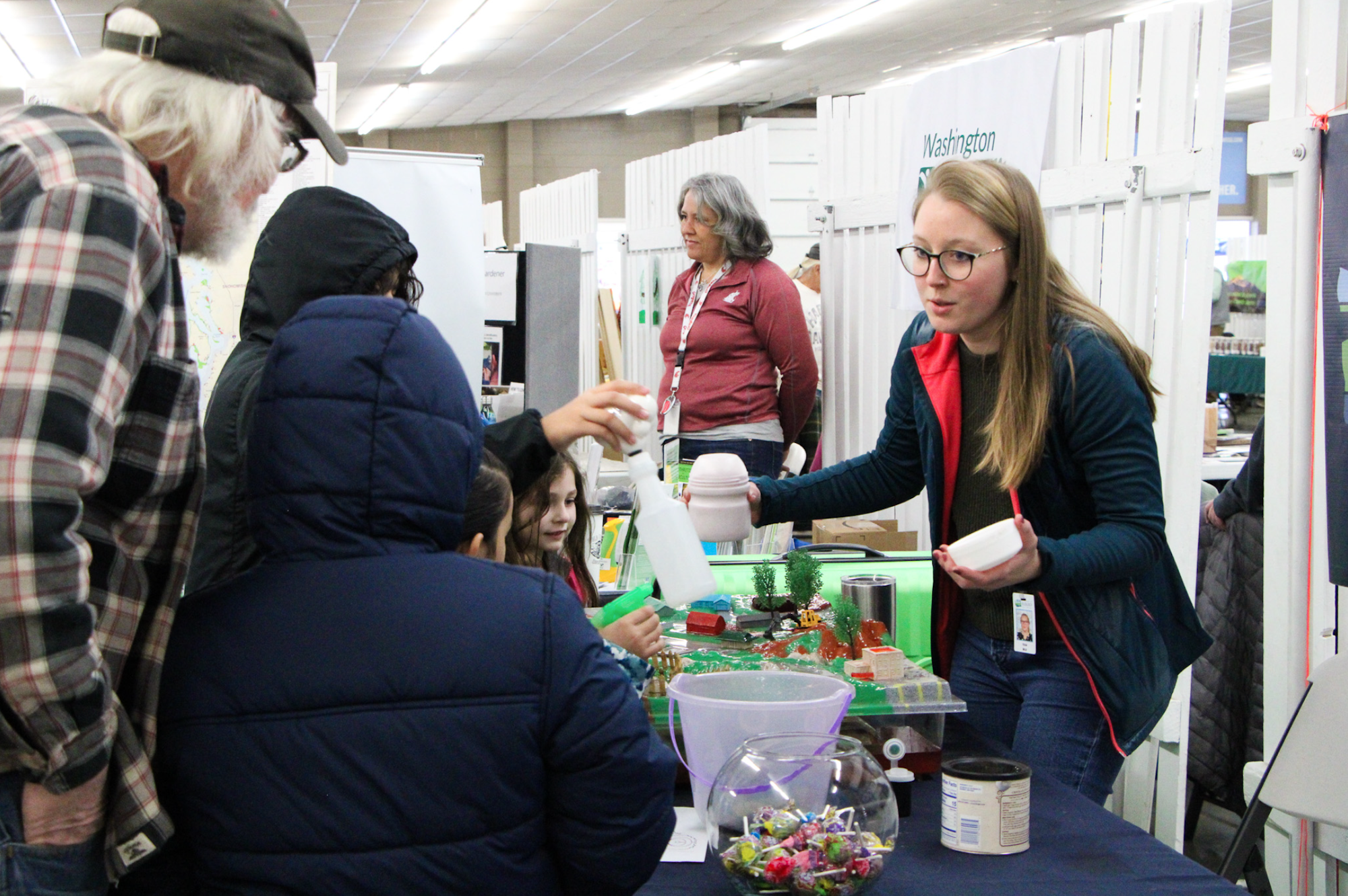 A group of people at a booth, interacting with a model display and containers, possibly in an educational or exhibition setting.
