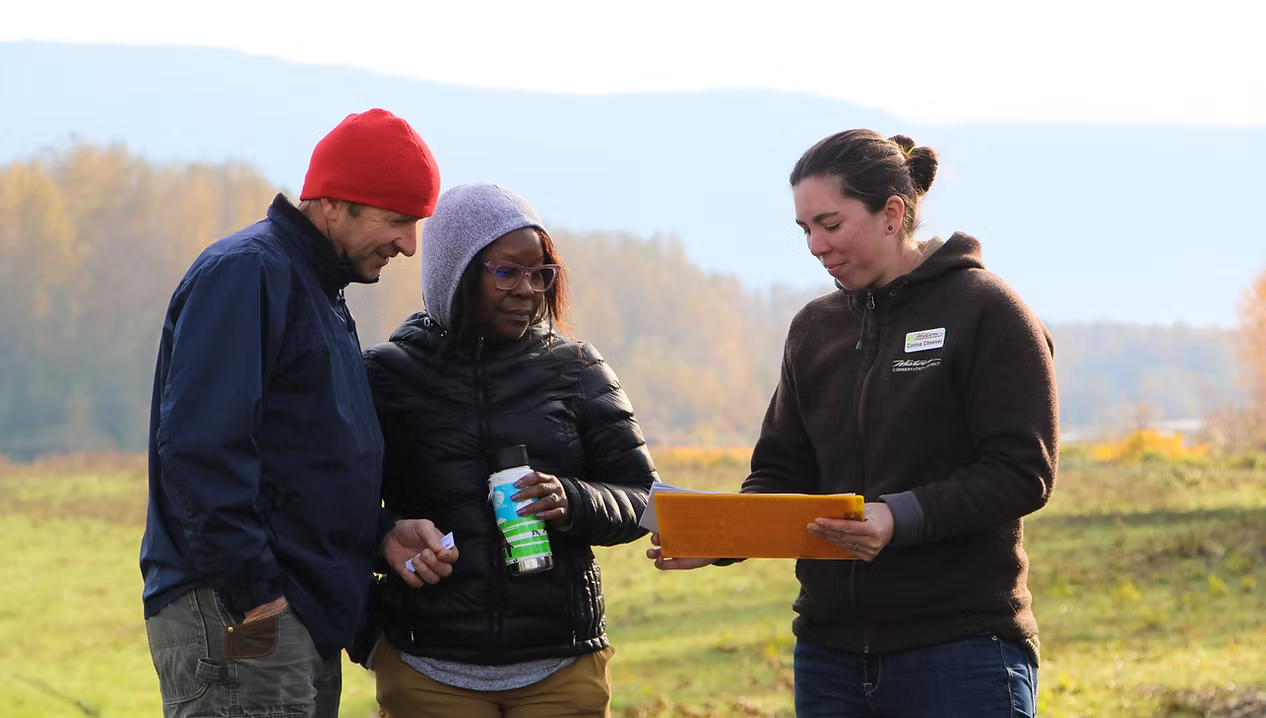 Three people outdoors, one holding a pad or clipboard, discussing something together.