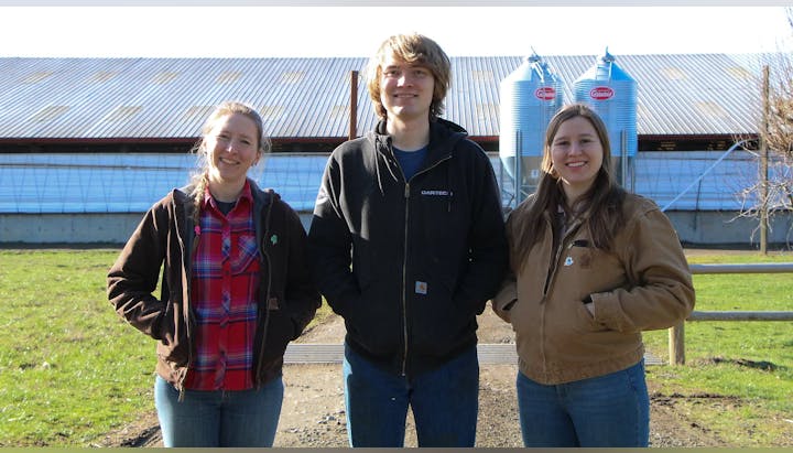Three people standing outside, smiling, in front of a building with silos.