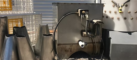 A metal industrial machine or equipment with hoses and pipes, surrounded by metallic cones and crates in a gravel area.