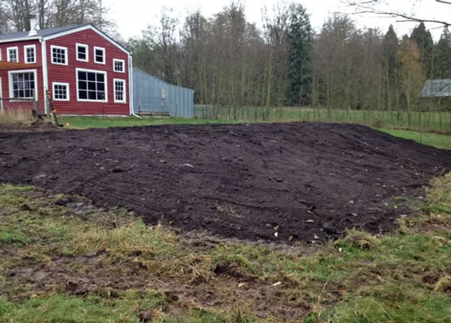 A red barn-like structure beside a freshly tilled field, surrounded by trees and grass.