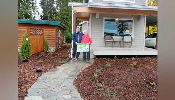 Two people standing on a walkway, holding a "We love our native plant garden" sign in front of a house.