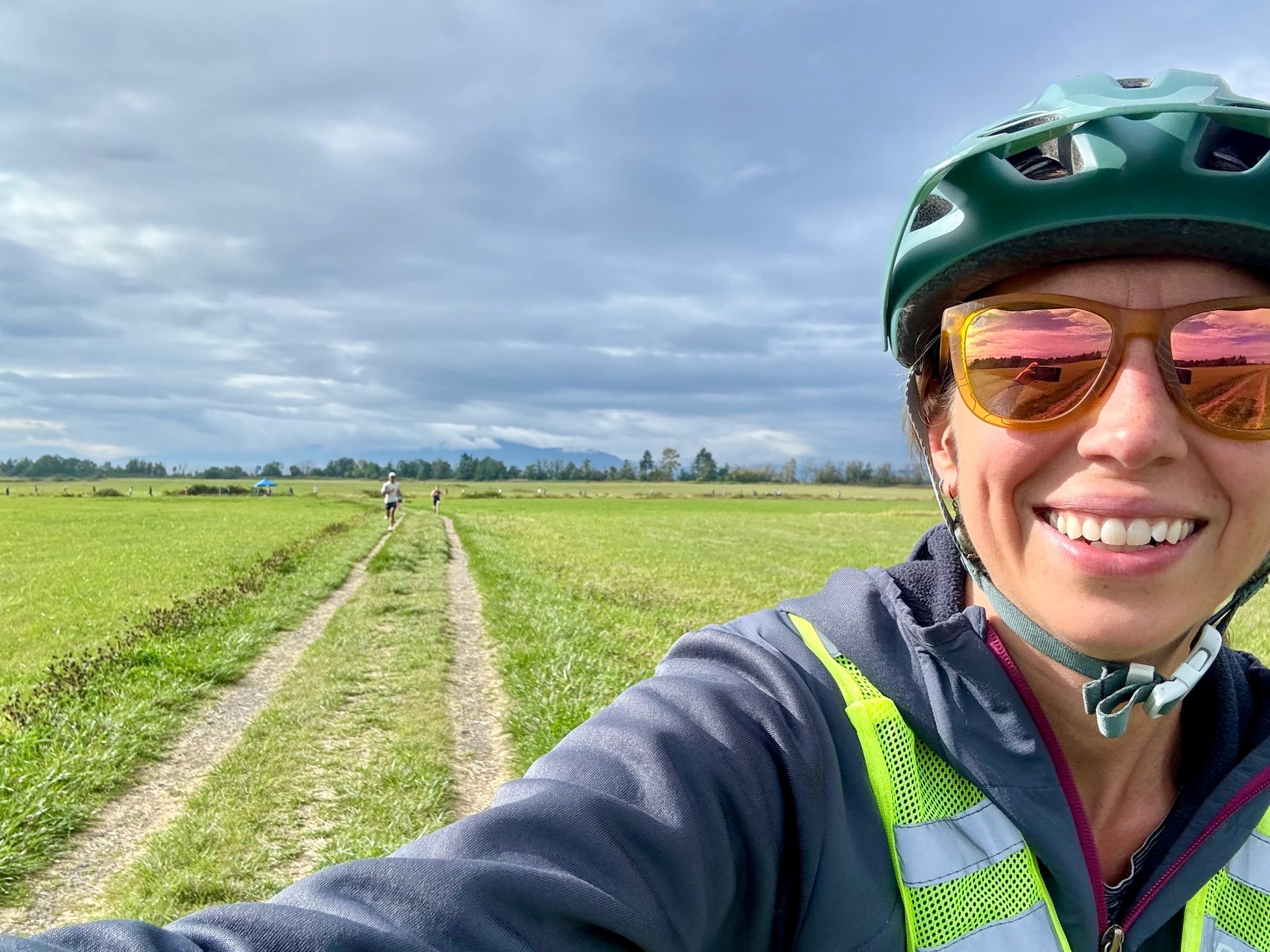 A person wearing a helmet and sunglasses is taking a selfie on a grassy path, with runners in the background.