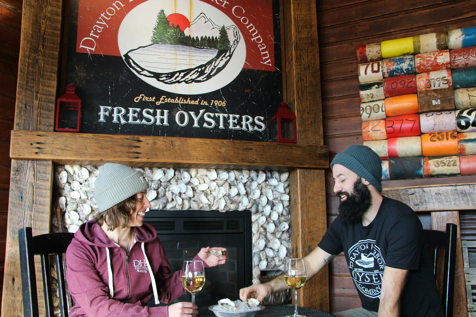 Two people enjoying fresh oysters and wine by a fireplace with oyster shells, under a sign for Drayton Harbor Oyster Company.