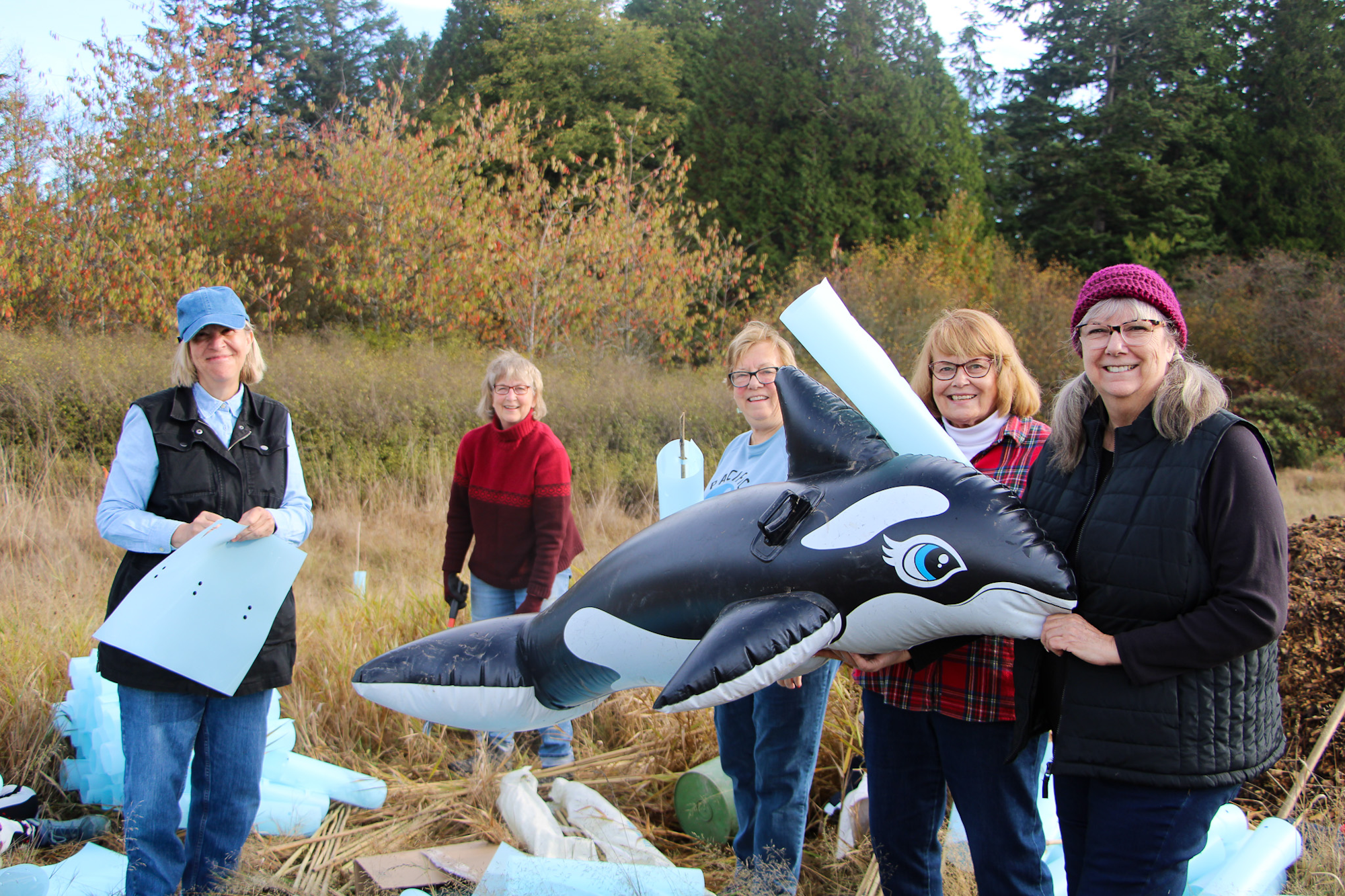Five people outdoors holding a large inflatable whale, surrounded by trees and grass.