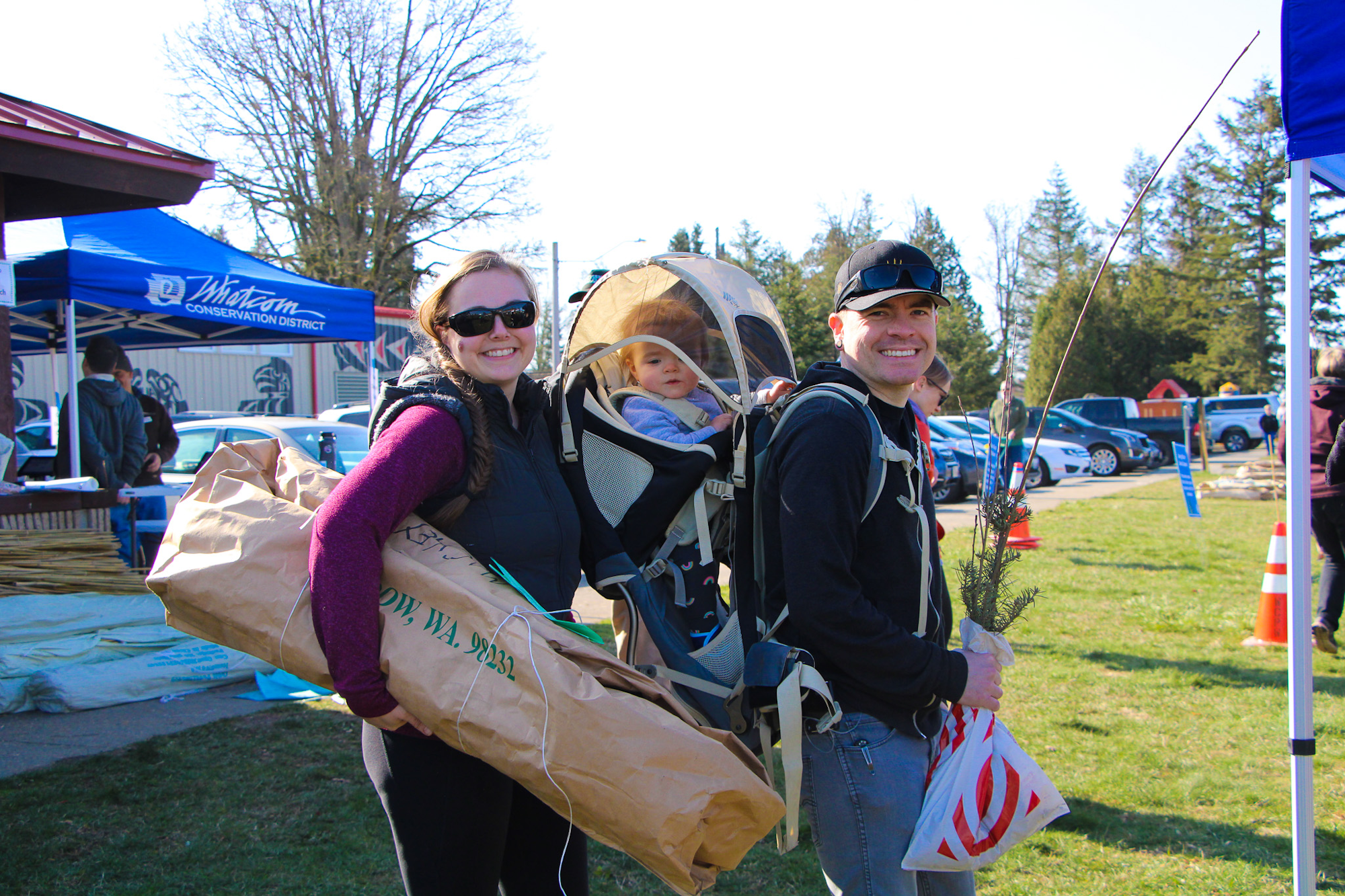A smiling family outdoors with a baby in a carrier, holding large packages, possibly at an event under a blue tent.
