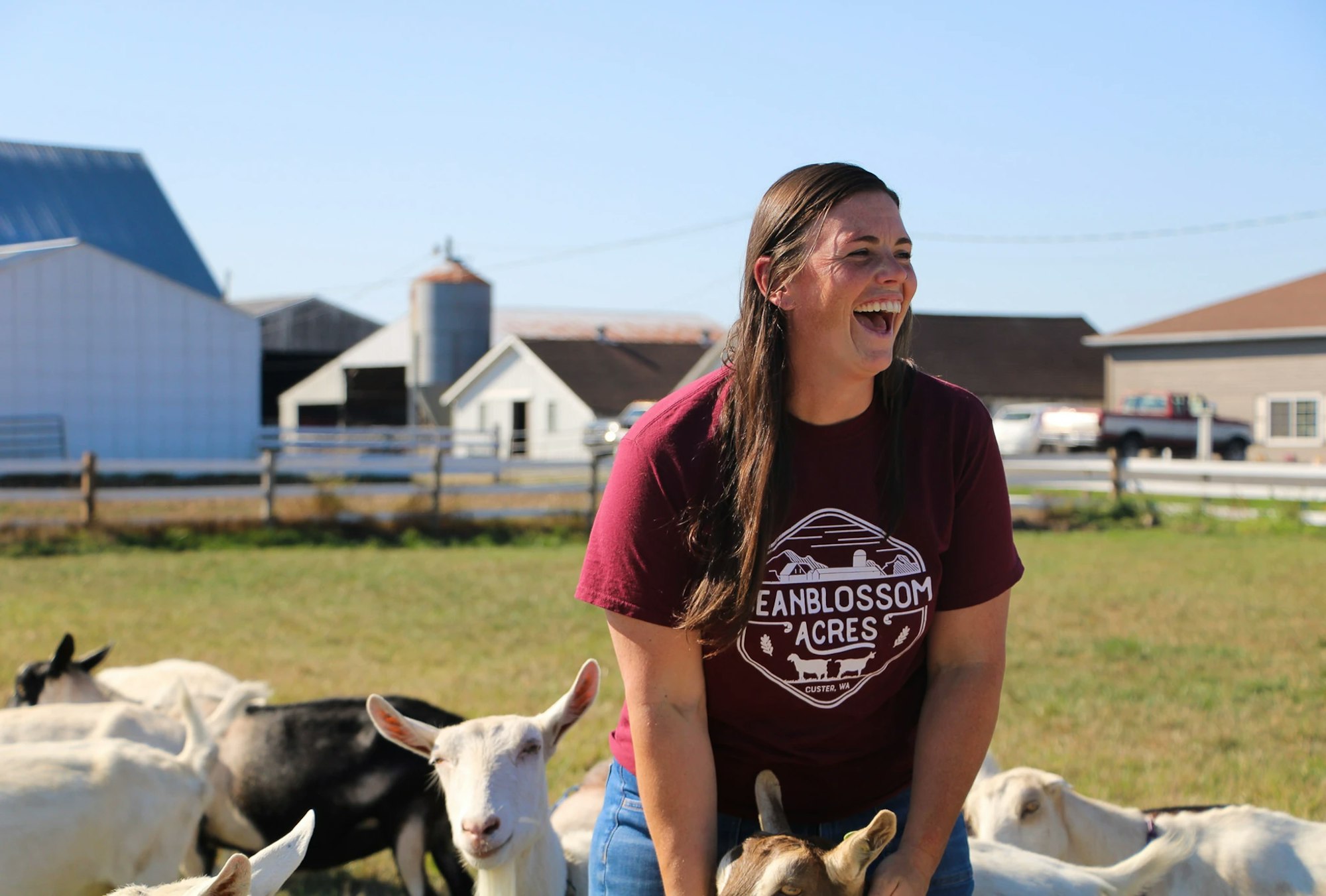 A person smiling with goats on a sunny farm.