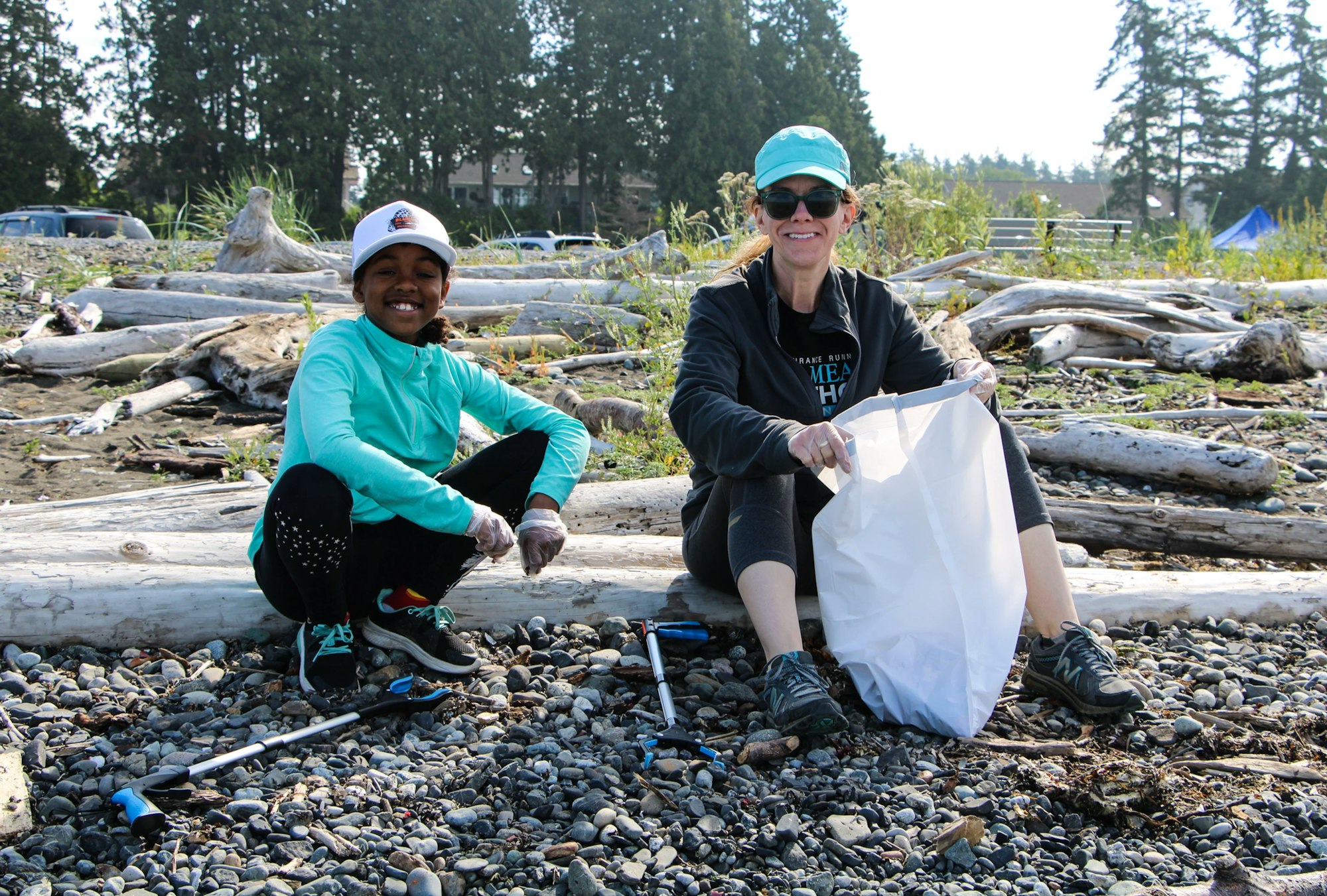 Two people are sitting on a rocky beach with trash bags, participating in a cleanup activity.