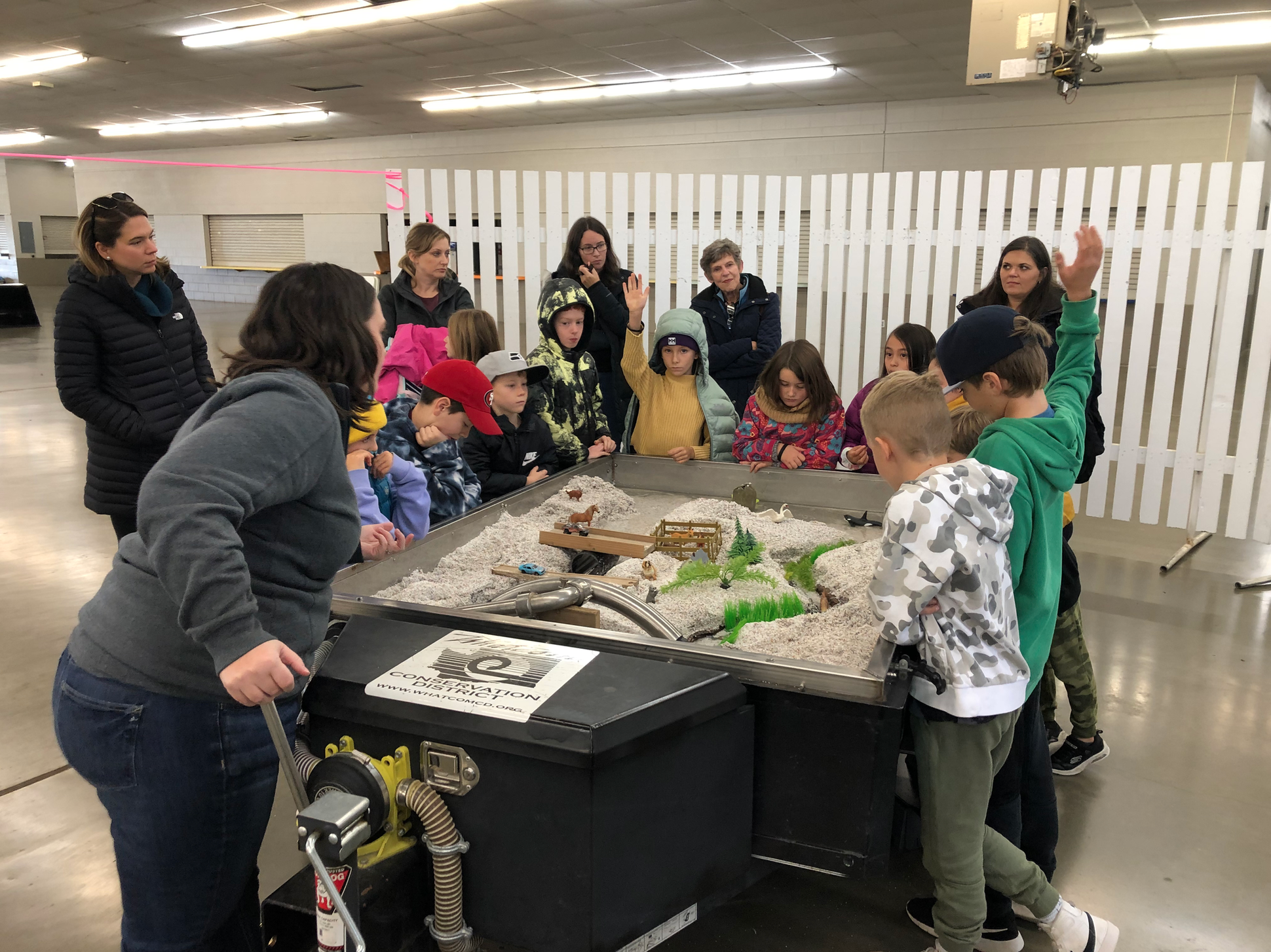 A group of children and adults gather around a model landscape display in an indoor setting.