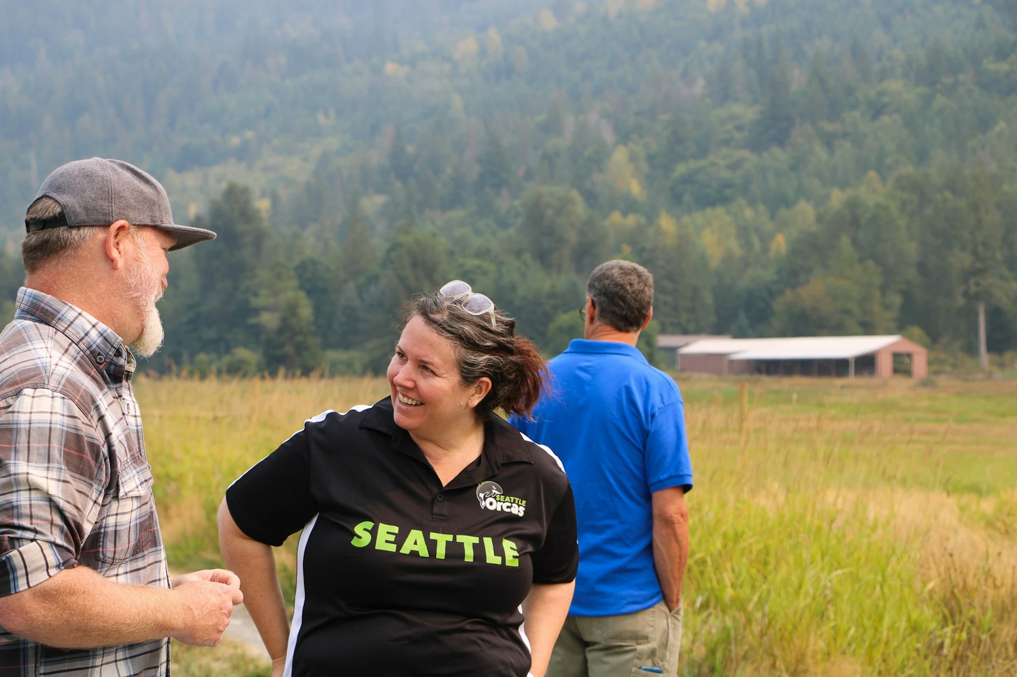 Three people outdoors, one woman smiling, two men standing, with fields and trees in the background.