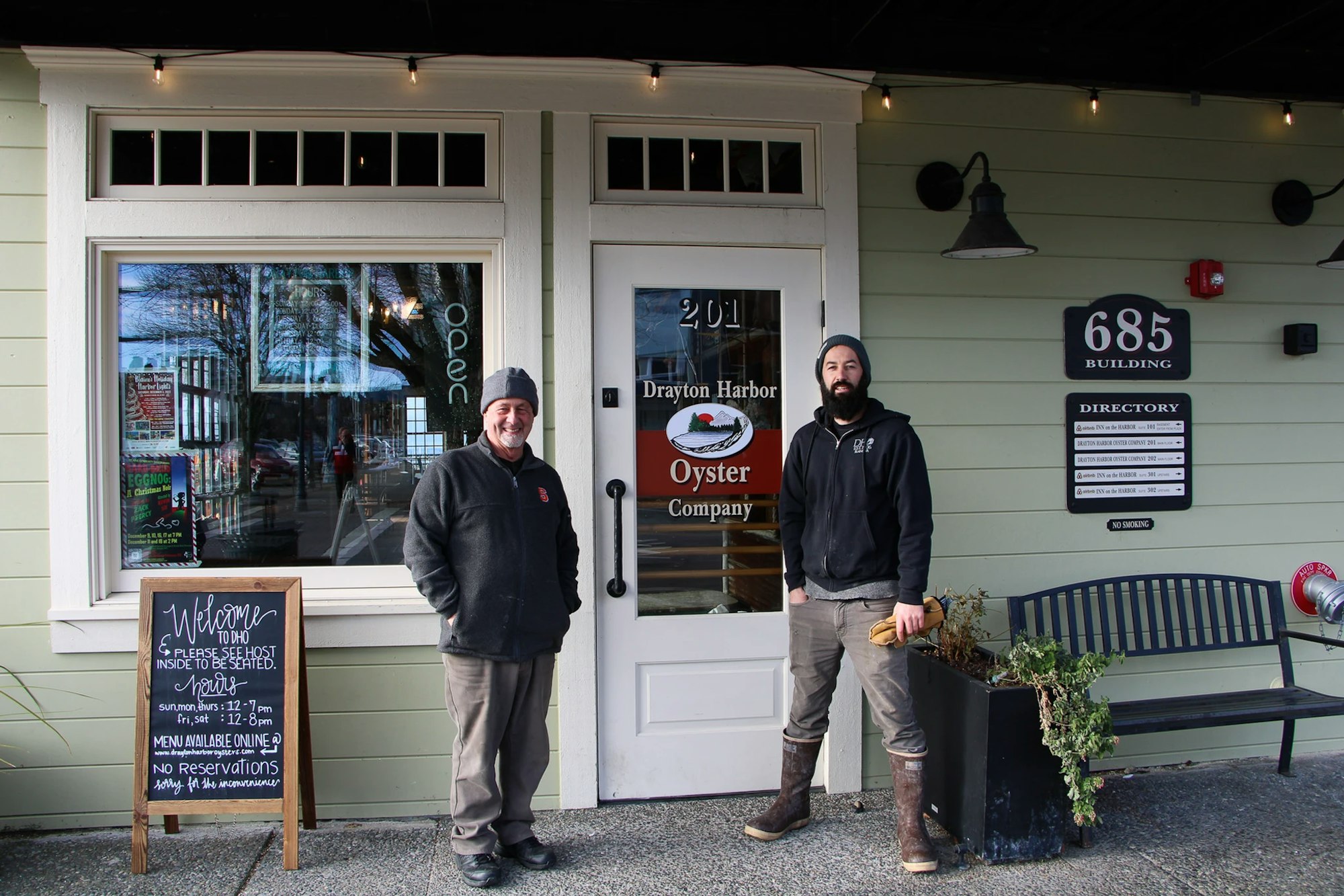 Two people stand outside Drayton Harbor Oyster Company, next to an "open" sign and a chalkboard with hours.