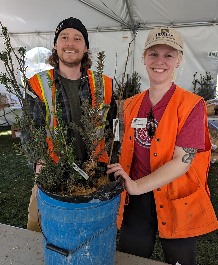 Two people wearing orange vests smile while holding a bucket with young trees inside a tent.