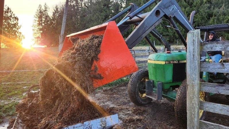 A tractor with a red front loader dumps dirt or compost on the ground with the sun setting in the background.