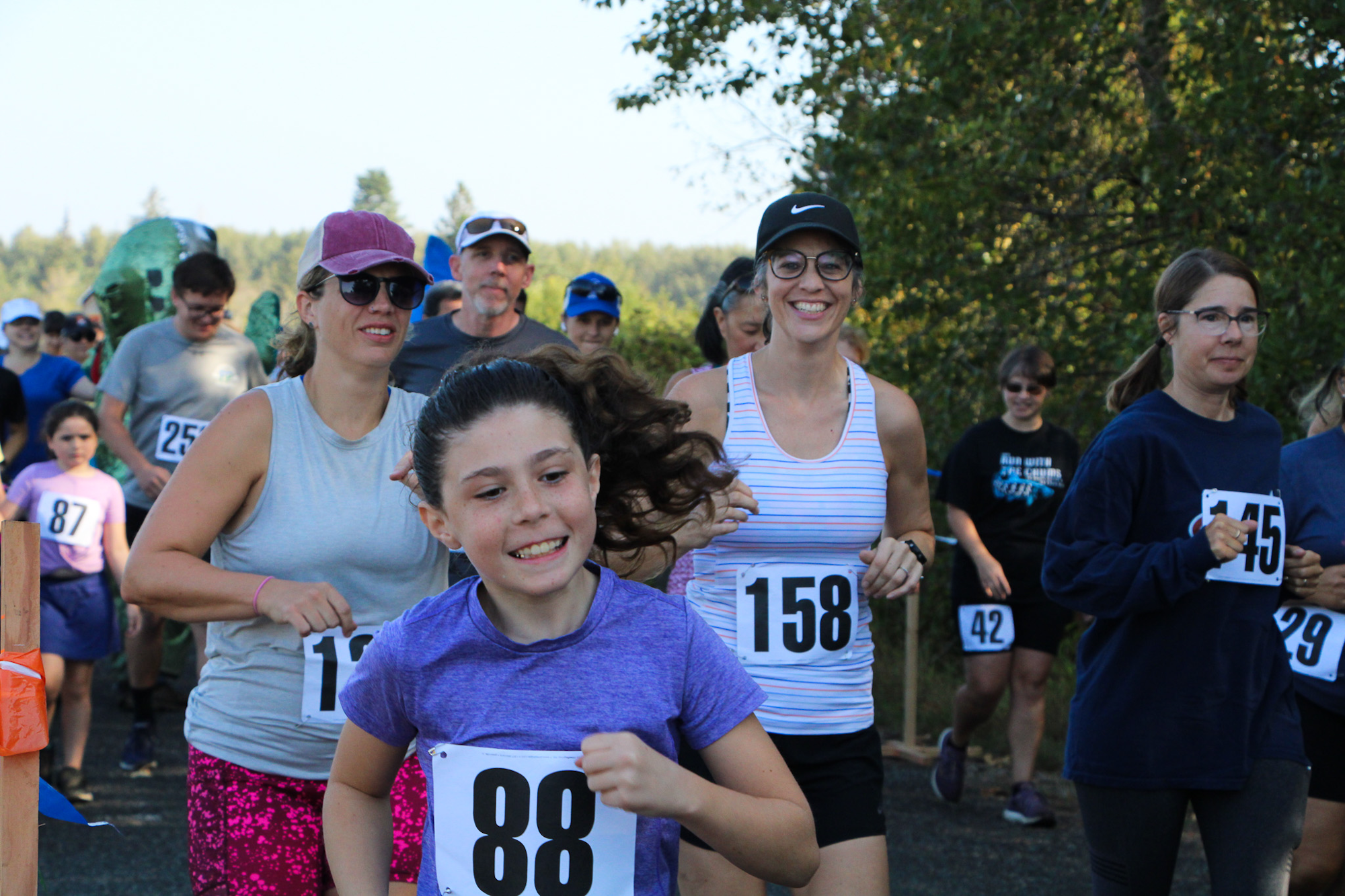 People running in an outdoor race, wearing numbered bibs, with trees in the background.